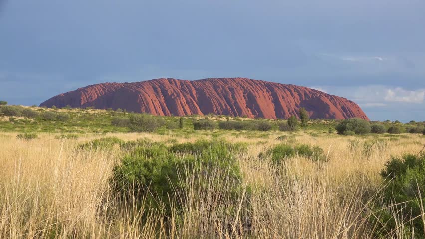 Iconic Australian outback monolith Uluru Ayers Rock under cloudy sky, natural desert landscape with native Australian plants grass and bushes.