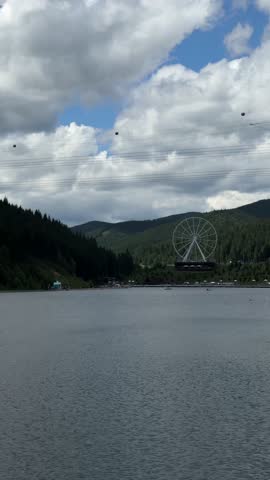A wide angle view of a large amusement park ferris wheel situated among dense green trees on a hill across a vast lake. Clouds drift slowly over the landscape under daylight.