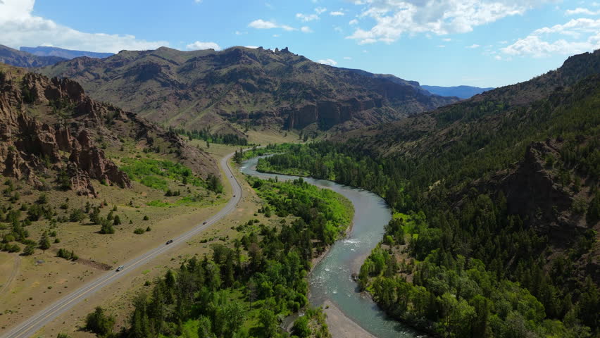 Aerial view of Montana river valley with turquoise water winding alongside scenic highway through rugged mountain canyon with lush green forests and dramatic rocky cliffs under bright blue sky.