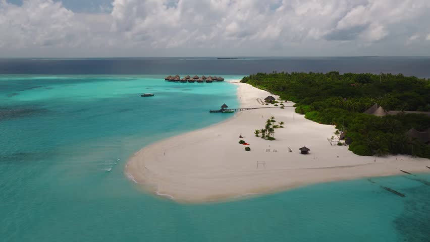 Aerial view shows a crescent island resort in Maldives, thatched overwater villas on a curved jetty, wide white sand beach, palm vegetation, and boats near the villas.