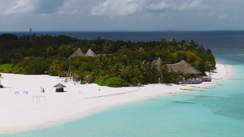 Aerial view of a crescent beach and turquoise lagoon in the Maldives, with thatched pavilions, a wooden jetty, a moored speedboat, orange kayaks, and two communication towers.