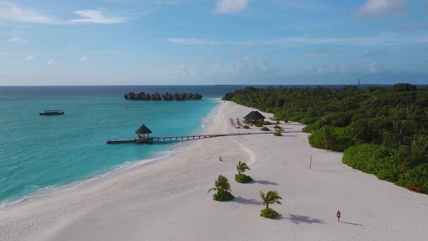 Aerial Maldives atoll shows palm sand crescent, wooden jetty, thatched gazebo, overwater villas, moored dhoni, tiny island on horizon, gentle waves, bright daylight.