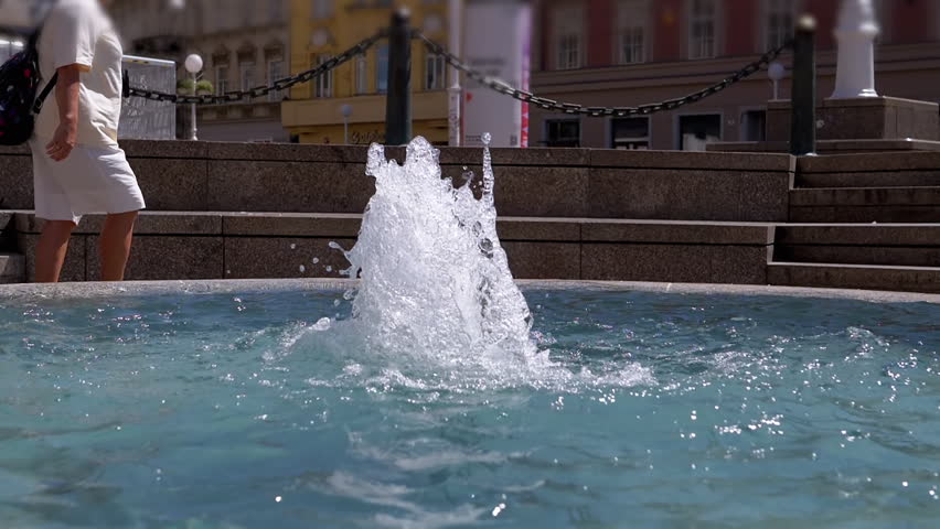 A small city fountain sprays water upwards as a person walks by in the blurred background. The scene evokes everyday life in an urban setting, ideal for projects about cityscapes and