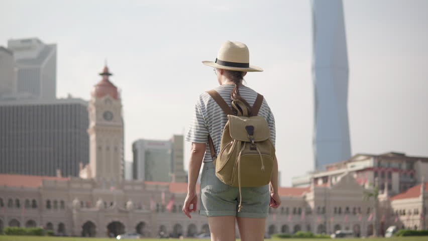 Female tourist in hat with backpack is raising her arms and enjoying view of the Sultan Abdul Samad Building and Merdeka 118 Tower from Independence Square (Dataran Merdeka) in Kuala Lumpur, Malaysia.