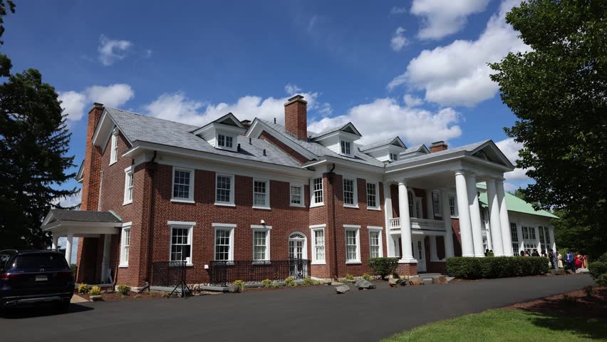 Large red brick mansion with white columns and a paved driveway under a blue sky with clouds. Elegant historic house exterior in a quiet leafy setting.