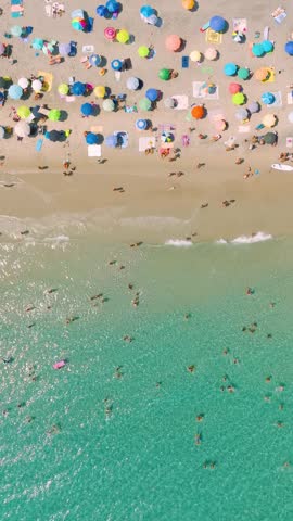 Vertical video. Top down aerial drone view of a crowded beach with colorful umbrellas and people relaxing. Crystal clear sea water and white sand during a bright sunny summer vacation.