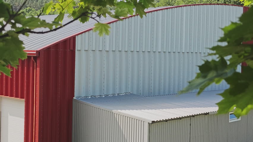 Modern red and grey metal farm building with a curved roof seen through green tree leaves on a sunny day