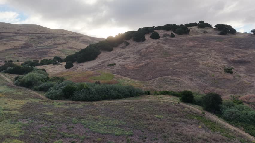 Cinematic aerial drone footage pushing forward over golden, oak-dotted hills near American Canyon, Napa County, California at sunrise. Soft morning light breaks through an overcast sky as the camera reveals rolling grassland, scattered oak groves, and a small rural barn below. Dry summer grass, tranquil ranch country, and layered foothills make this ideal for nature, travel, and California landscape projects. 4K, no people.