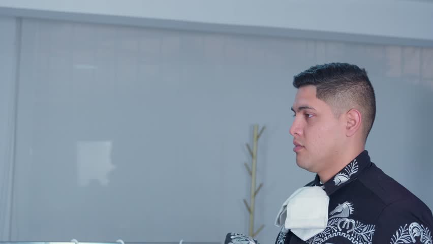 Side profile of a young hispanic man in a traditional charro suit putting on an embroidered sombrero hat before his performance