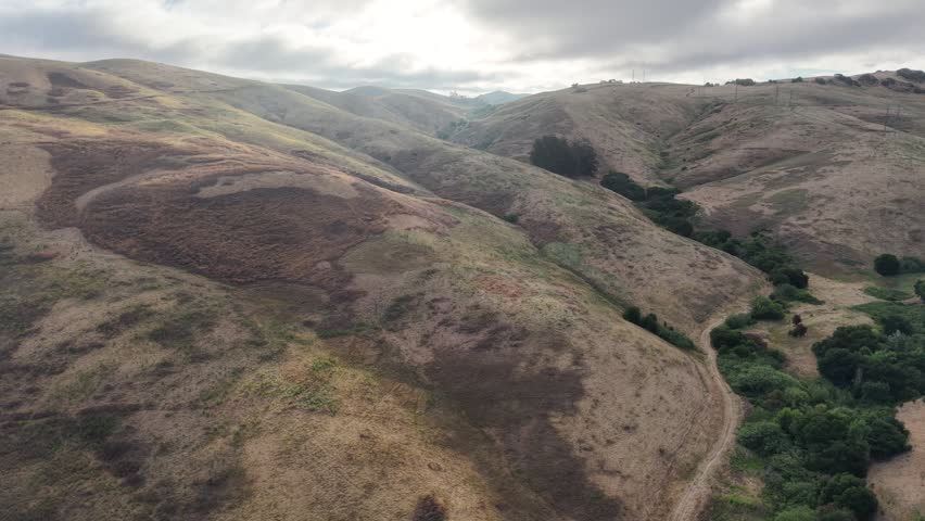Aerial Over Golden Hills and Creek Drainage Toward Peak, American Canyon, 4K. Cinematic 4K aerial drone push over golden rolling hills and a broad creek drainage near American Canyon, Napa County, California, beneath a soft overcast morning sky. A trail leads the eye toward a distant peak across layered grassland and green vegetation. Expansive and tranquil — ideal for nature, travel, hiking, and California landscape projects. No people in frame.