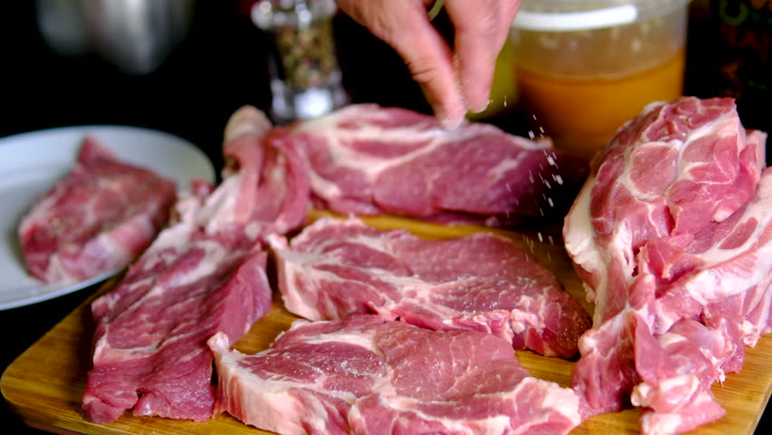 Male hand sprinkling coarse salt over pork neck steaks on wooden cutting board before cooking