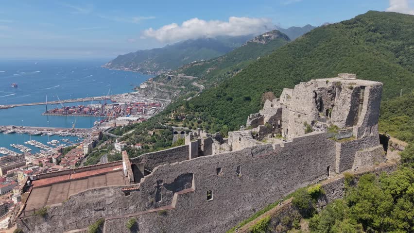 Aerial view of Castello di Arechi stone ruins and ramparts overlooking Salerno container port, marina, and Amalfi Coast mountains, Italy