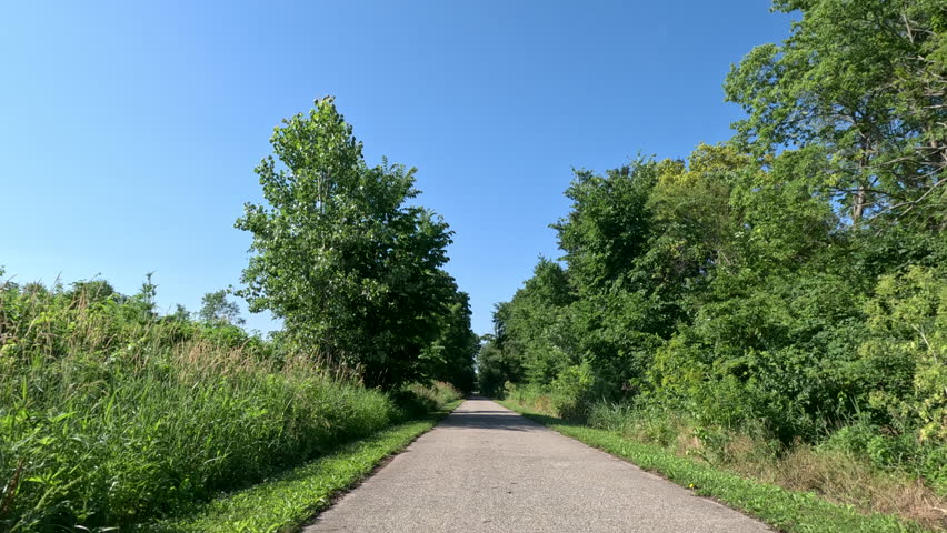 Paved Multi-Use Trail Passing Through Trees and Wild Grass. A smooth, slow-motion view from a moving bicycle receding down a paved recreational trail lined with lush green trees and tall wild grass under a clear blue sky.