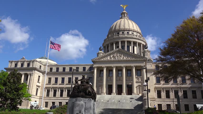 Wide shot of historic Mississippi state capitol building. Landmark architecture features grand dome and columns. American flag flies in breeze against blue sky. Static low angle clip.