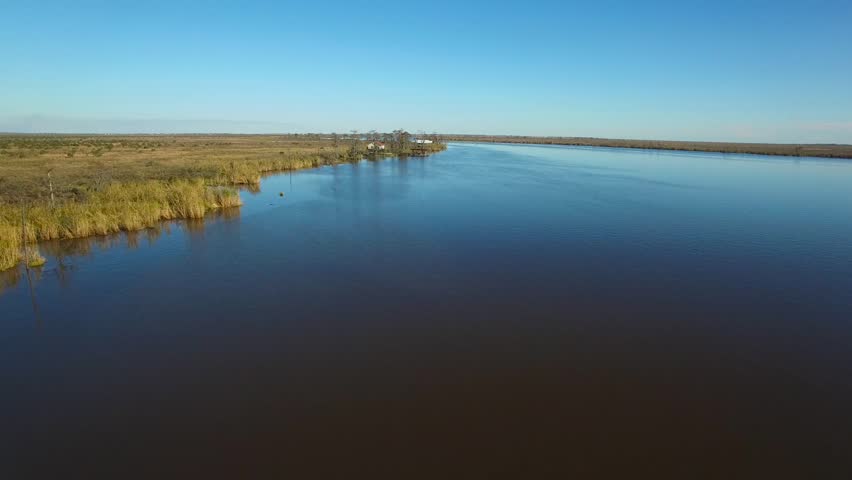 Aerial video drone flying over vast Louisiana bayou landscape. Moving towards small group of houses sitting on bayou river bank. Houses are surrounded by cypress trees and green marsh grass. The water reflects blue sky.