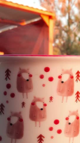 Patterned mug beside red outdoor shelter in warm sunlight, with repeated sheep illustration and red dots visible, while background trees softly blur and light shifts across mug surface