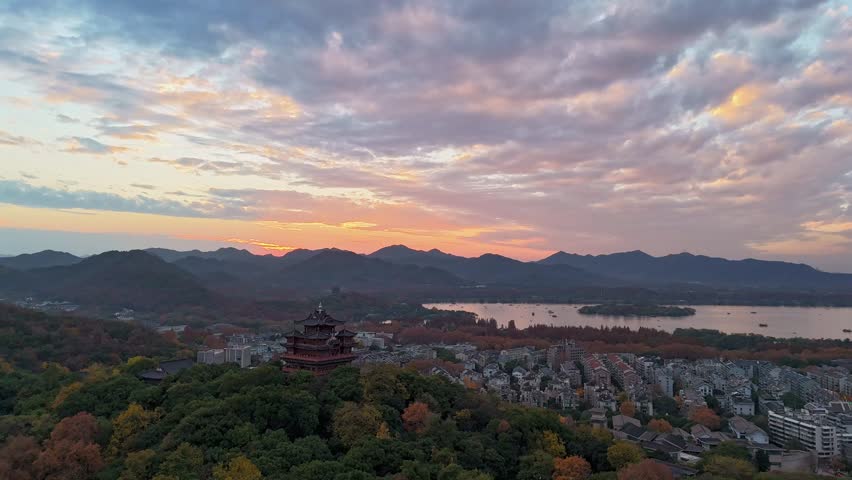 Aerial view of Wu Hill with the historic City God Pavilion at sunset, overlooking the iconic West Lake, Leifeng Pagoda, and distant mountains, capturing the stunning autumn foliage and vibrant sunset sky.