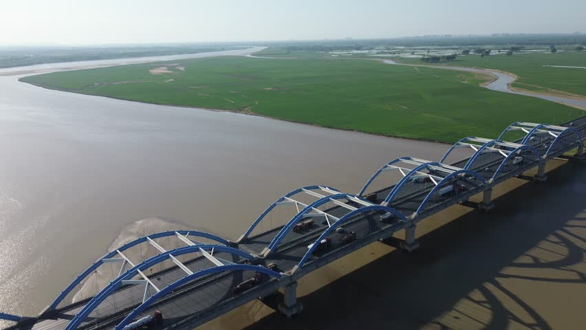 Cinematic forward-flying high oblique aerial view of iconic blue steel arch bridge spanning the vast Yellow River. Massive rhythmic arch shadows stretch across the silty riverbed, with endless green farmland and winding wetland waterways stretching to the horizon. Modern transportation engineering blends with majestic alluvial river landscape and lush rural scenery in bright clear daylight.