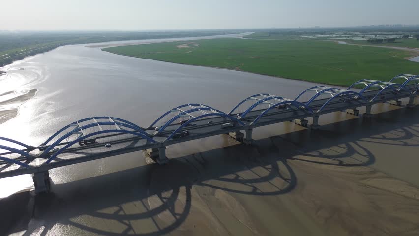 Pull-back high-angle aerial drone shot of Liujiang Yellow River Twin Steel Arch Bridge in Zhengzhou, Henan, China. The blue-white arched highway bridge stretches across the wide silty Yellow River, with giant repeating arch shadows cast on exposed alluvial sandbars. Lush green farmland and winding river floodplain spread across the far riverbank, mixed passenger and freight vehicles travel on eight-lane carriageway, bright sunny daylight, distant river bend and city skyline on the horizon.