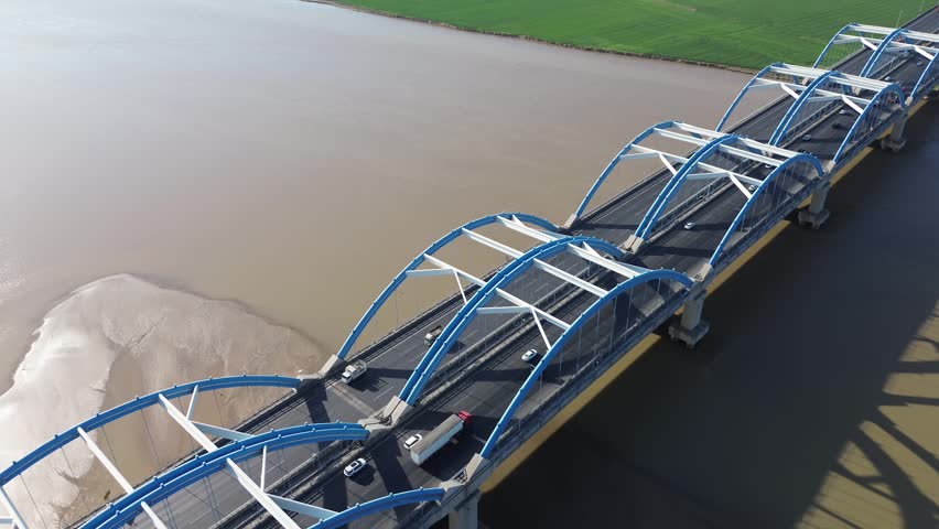 Cinematic forward-flying oblique aerial view of iconic blue steel arch bridge spanning the Yellow River. Massive curved arch shadows stretch dramatically across the silty river surface and sandbars, with steady traffic flowing along the elevated highway deck. Modern bridge engineering blends with vast natural river landscape and green farmland in bright clear daylight.