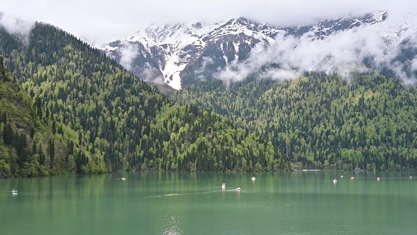 The emerald green waters of Lake Ritsa with small boats, framed by dense wooded hillsides and majestic snowy mountains, Abkhazia
