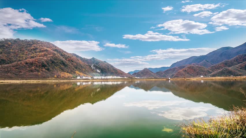 Time lapse captures wide lake surrounded by autumn color forest,warm tone clouds drift slowly across sky,layered color mountain fully reflect on gentle lake water