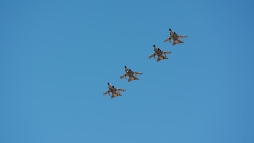 Four camouflaged military jets perform a coordinated aerial display, flying in tight diagonal formation against a clear blue sky, captured in slow motion