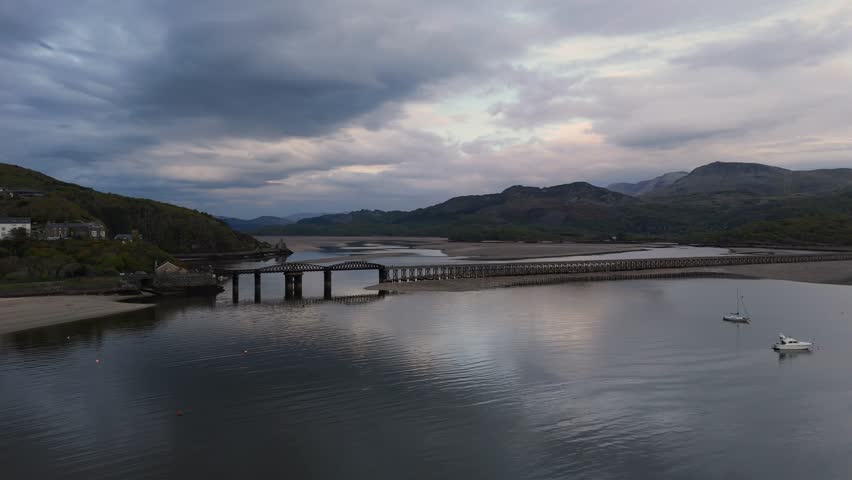 A drone view of Barmouth Bridge crossing the Mawddach Estuary with boats, surrounded by mountains in North Wales, UK. During sunset