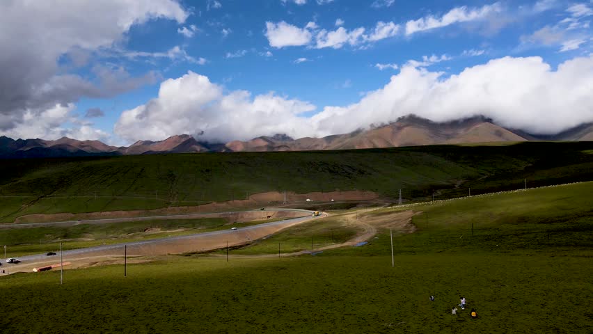 Drone overlooks vast plateau meadow of Qinghai China,straight highway crosses whole green grassland,large white clouds drift slowly above continuous distant mountains