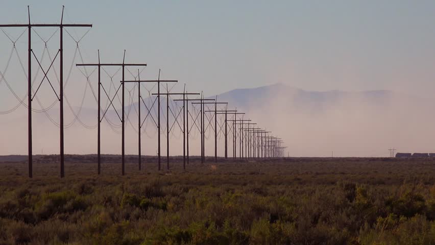 A long line of electricity pylons recedes into the distance, with mountains shrouded in haze behind. Desert landscape with dry brush.
