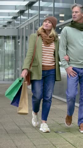 Vertical video: Leaving shops, walking couple arm-in-arm in coats, carrying shopping bags. Romance, companionship, stroll, arcade, canopy, outdoor, vertical video