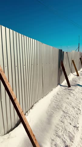 Snow-covered corrugated steel fence leaning on wooden braces, winter construction site under bright blue sky, deep snowdrifts piled