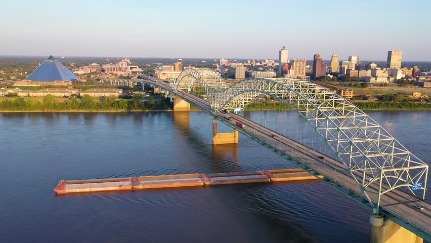 Dynamic aerial footage shows barge moving along Mississippi River beneath iconic Hernando de Soto Bridge at sunset with Memphis city skyline and pyramid landmark in background during golden hour.