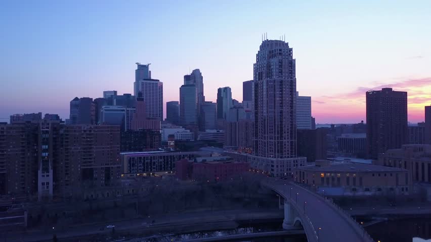 Aerial view of downtown minneapolis skyline at dusk, showcasing modern architecture and a prominent bridge over the mississippi river with traffic motion during evening hours against colorful sky.