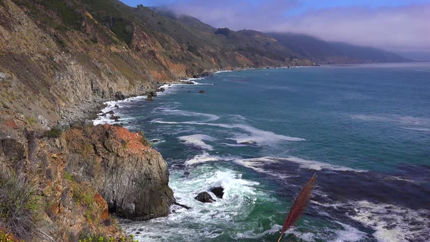 Dynamic ocean waves crash against a rugged California coastline featuring rocky cliffs and lush green mountains under a bright sky with misty clouds, creating a dramatic and serene natural landscape.
