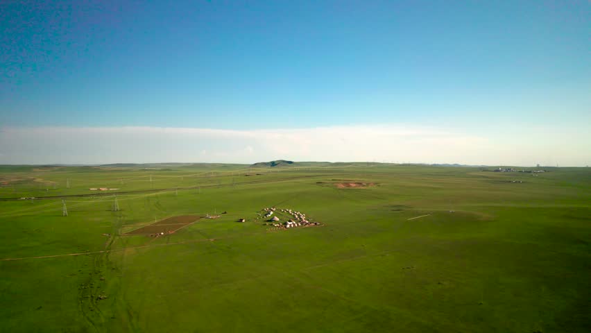 Drone overlooks wide flat green grassland Inner Mongolia China,small yurt settlement sits in center of meadow,fluffy white clouds drift slowly over low hills