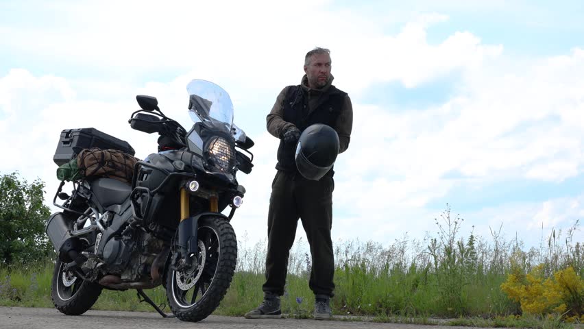 A middle-aged motorcyclist puts on a helmet, mounts his touring motorcycle, and rides off. Long motorcycle rides serve as a hobby and an escape from the information noise of big cities.