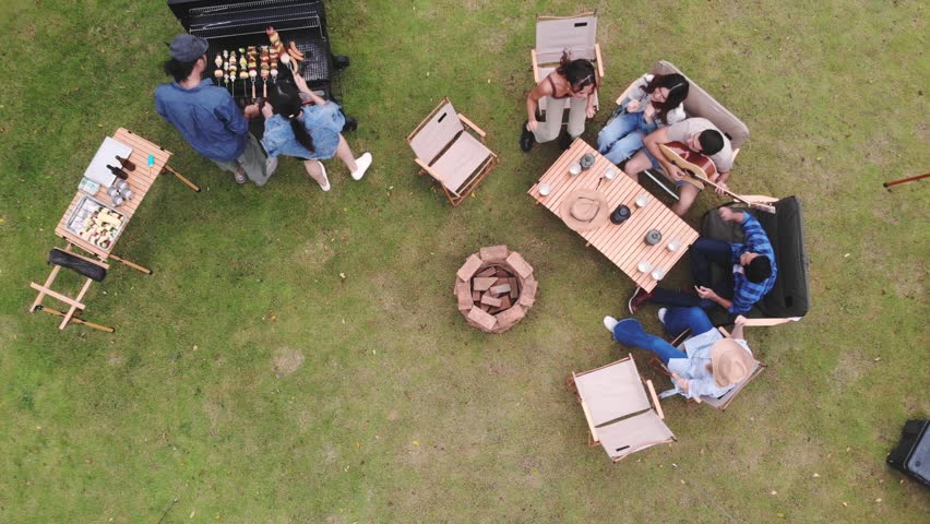 Aerial top-down view of a group of friends enjoying an outdoor barbecue party and camping on a green lawn, featuring a charcoal grill, wooden dining table, chairs, and a fire pit