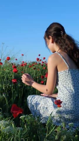 Girl crouching to touch poppies in a flower field under a clear blue sky