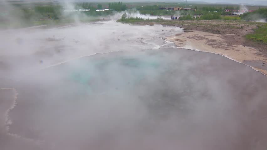 Dynamic overhead shot shows thick white steam rising from turquoise hot spring. Bubbles emerge from thermal water while people observe natural volcanic activity on barren landscape of Geysir park.