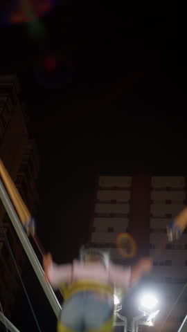 Child leaping high into the night air on a bungee trampoline at an amusement park, held by stretched elastic cords and a harness, floodlights and apartment towers below