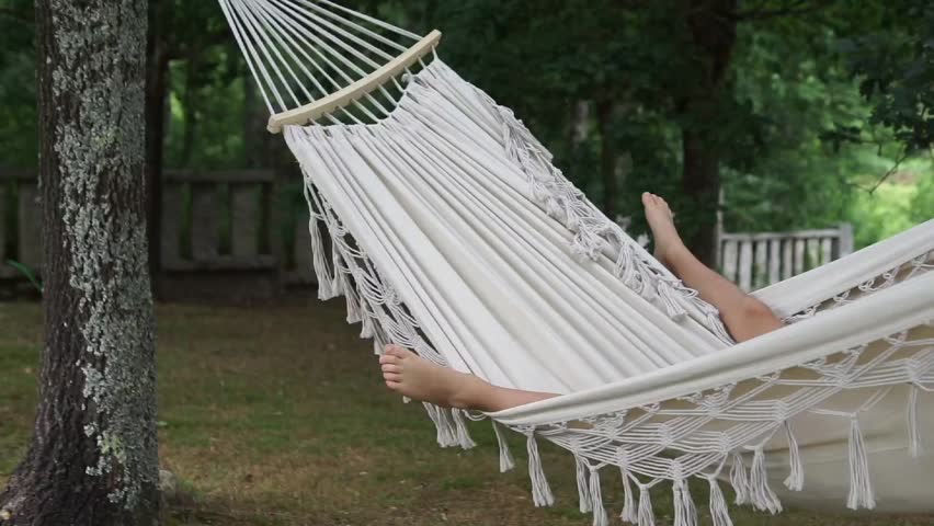 Wide tracking shot of a white fringe hammock hanging between trees in a big backyard, swinging slowly while a person relaxes inside during summer break.