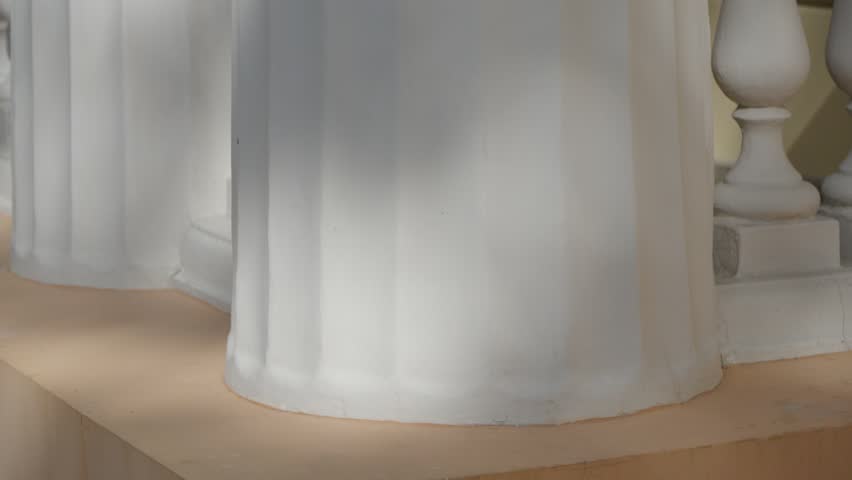 A slow tilt up captures the close up details of fluted white columns and a stone balustrade on the porch of a classical building facade.