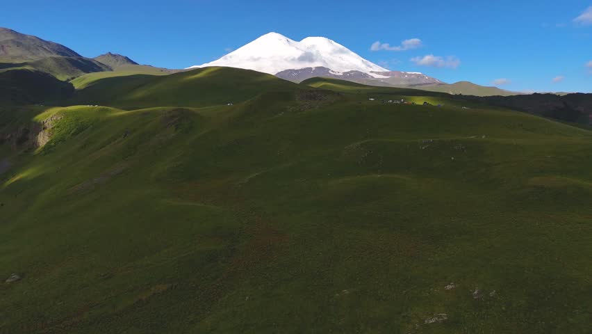 Drone view of the snow-capped peaks of Mount Elbrus and its green slopes at sunrise, with clouds in the sky