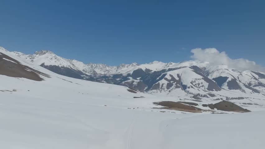 Drone footage of snow covered mountain landscape with a winding road and wide valley under blue sky in Grigoryev Gorge, Issyk Kul Region, Kyrgyzstan