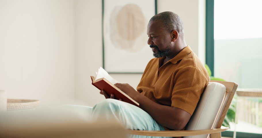 Relax, black man and reading book in house with funny story, smile and literature on weekend break. Novel, happy and mature person on armchair at living room with fantasy humor or laughing in home