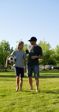 Father and son walk through park at sunset, smiling and discussing soccer game after playing together. Dad embraces child while boy carries soccer ball in vertical video celebrating family connection.