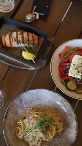 Vertical screen Restaurant table with grilled salmon creamy pasta and fresh Greek salad showcasing balanced healthy lunch Mediterranean cuisine and dining experience