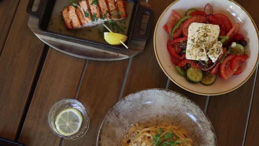Top view of grilled salmon Greek salad creamy pasta and lemon water served on a restaurant table highlighting healthy Mediterranean dining and balanced meal choices