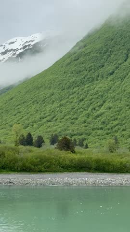 Snow-covered mountains rising above lush green hills with low clouds drifting across the landscape in Disenchantment Bay, Alaska.
